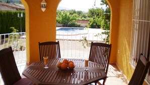 A wooden table with chairs, a glass of juice, and a bowl of oranges on a balcony with a view of a swimming pool and greenery.