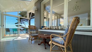 A sunlit room with a view of the sea, featuring wicker furniture, a wooden table, and a window with decorative jars.