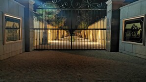 Un ingresso recintato con un cancello in ferro battuto decorativo, illuminato da luci soprastanti, incorniciato da due cartelli con la scritta "Quinta Vita".