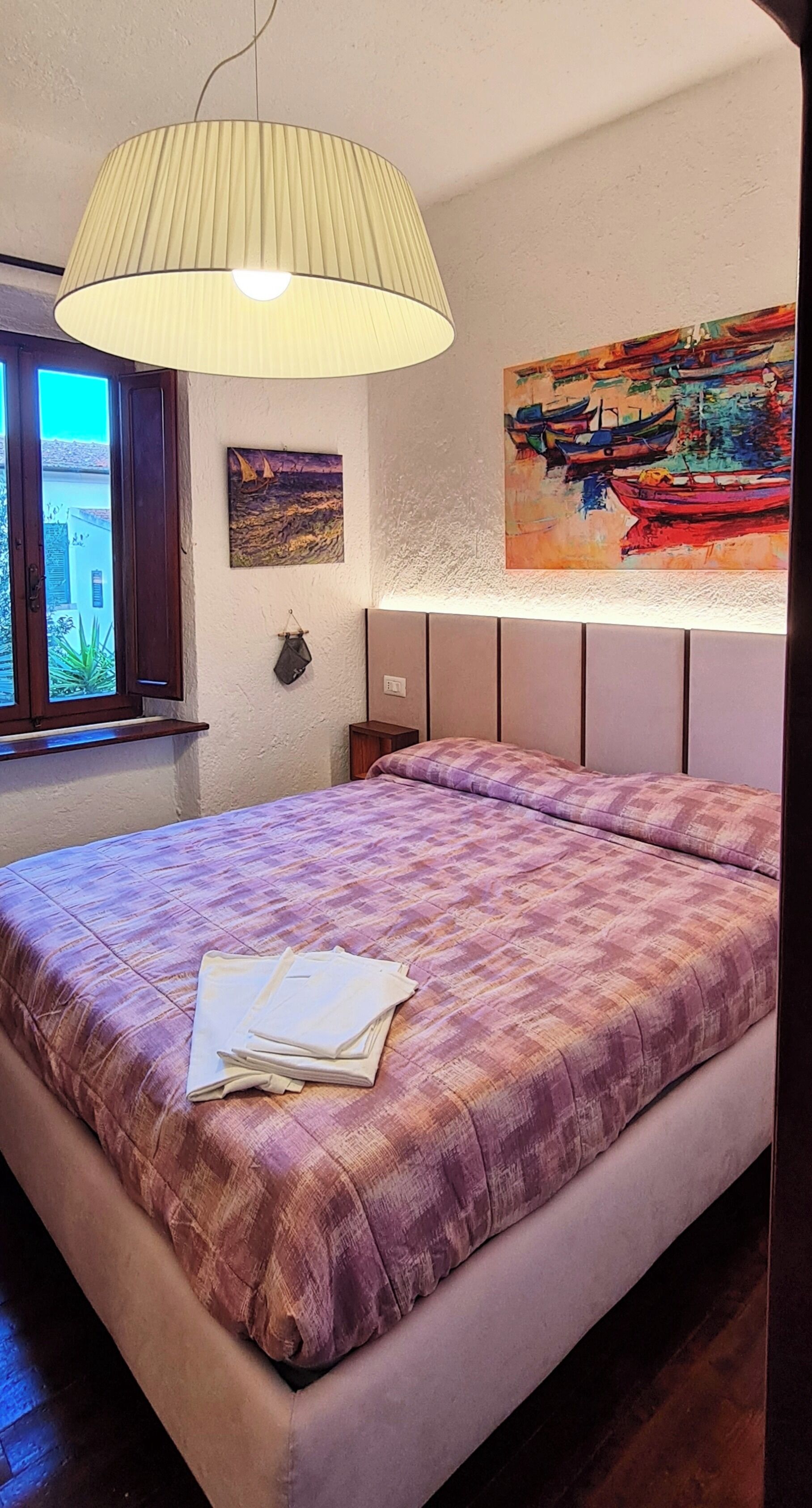 Ein Schlafzimmer mit einem Bett, einem Fenster, einem Gemälde und einer Pendelleuchte.
