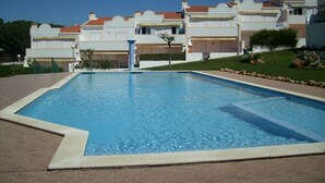 En swimmingpool med fliseterrasse, omgivet af en anlagt have og en række bygninger med altaner.