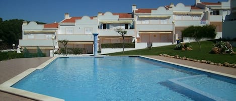 En swimmingpool med ett kaklat poolområde, omgiven av en vacker trädgård och en rad byggnader med balkonger.