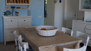 A dining area with a wooden table and white chairs, a shelf with dishes and kitchenware, and a door leading to another room. - beautiful apartment in villa, private beach with umbrellas and sunbeds (Marzamemi)