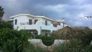A two-story white house with a flat roof, surrounded by lush greenery and a thatched structure. - beautiful apartment in villa, private beach with umbrellas and sunbeds (Marzamemi)