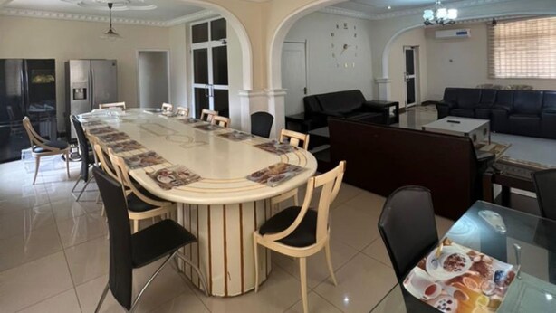 A spacious living area with a large round table, comfortable seating, a ceiling fan, and a modern kitchen in the background. - 6 BEDROOM HOUSE  IN COMMUNITY 20, GREATER ACCRA , TEMA OFF THE SPINTEX ROAD (Tema New Town)