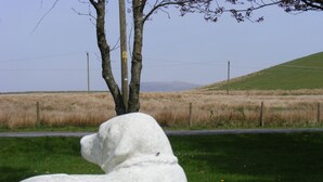 A white sculpture of a dog in a grassy area with a road and a tree in the background. - Traditional Stone Cottage (Ammanford)