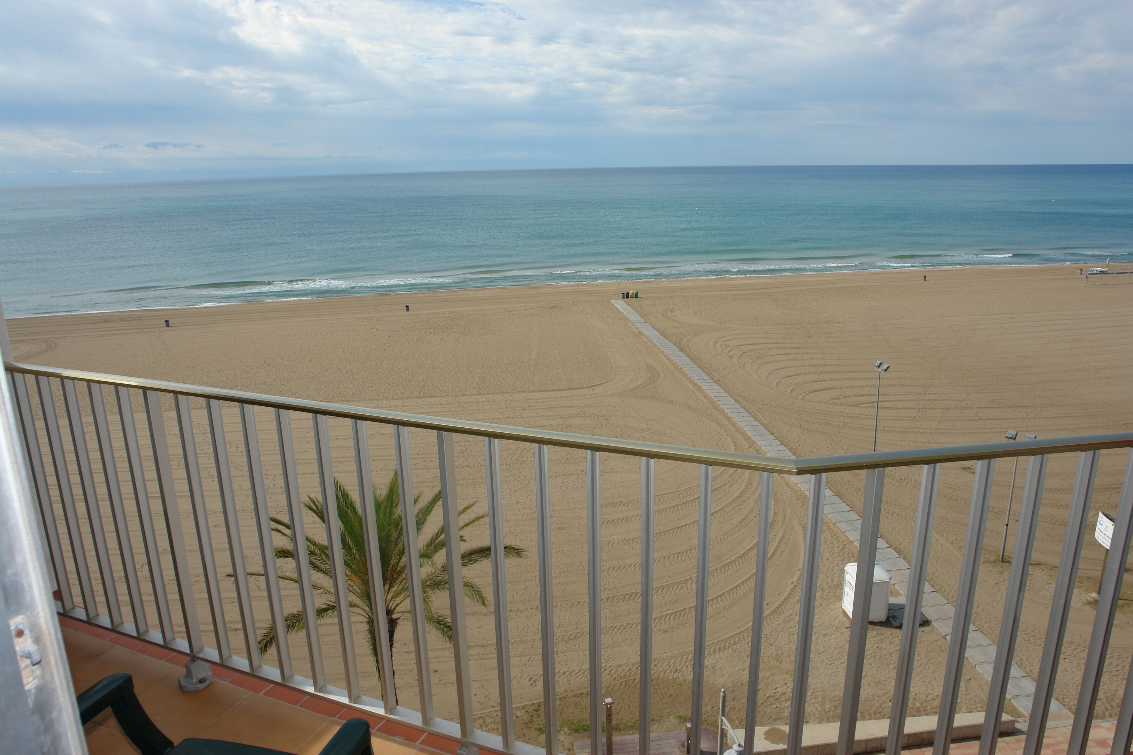 A beach view from a balcony with a railing, palm trees, and a clear sky.