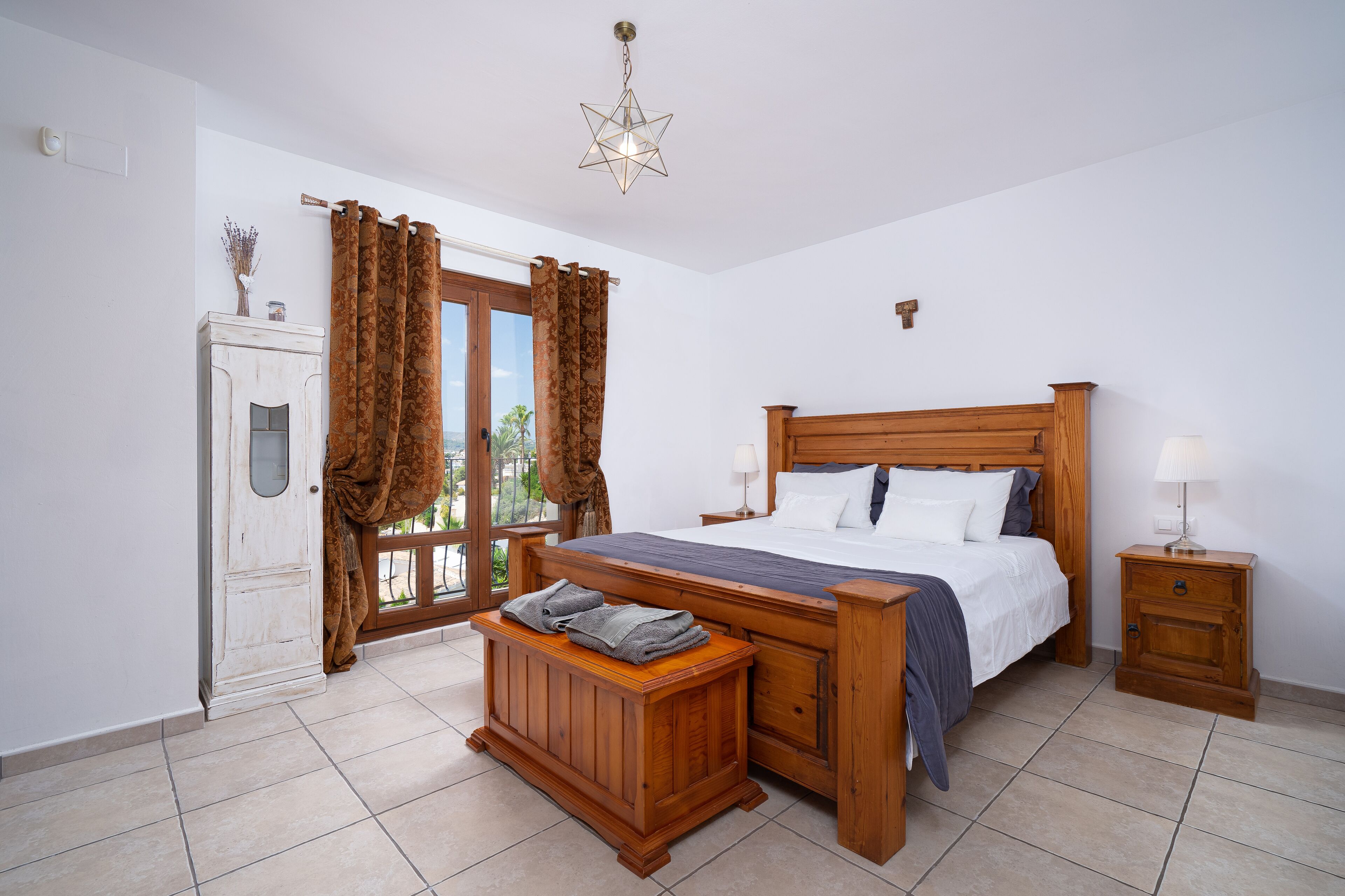 Een slaapkamer met een houten bed, nachtkastje, bankje, garderobe en een raam met gordijnen.