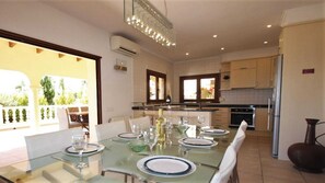 A modern kitchen with a dining area, featuring a glass table set for four, a built-in oven, and a view of the outdoors. - Moraira Sunset Villa with Private Pool, Sleeps 10, Family Friendly (MORAIRA)