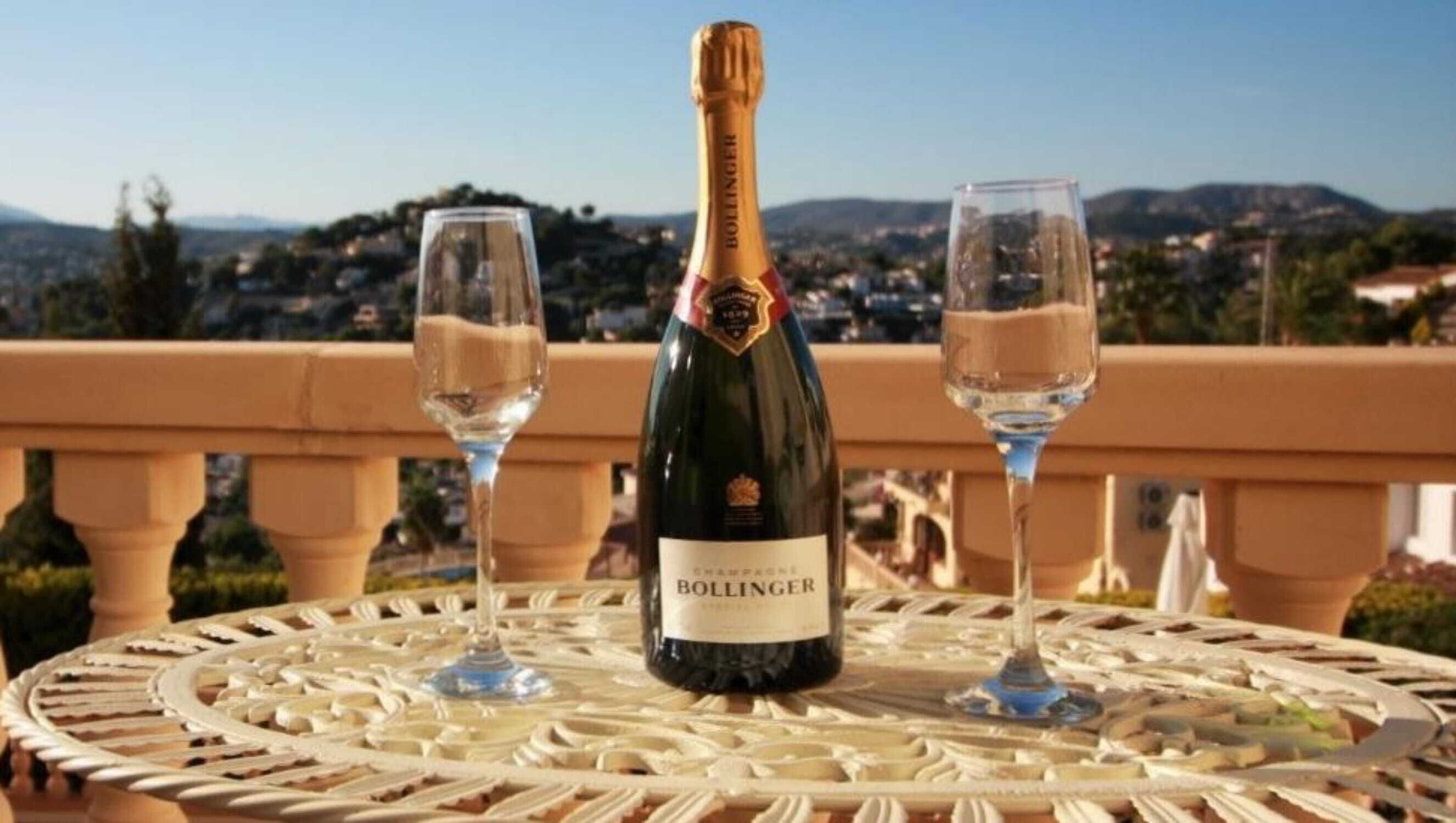 Una botella de champán Bollinger y dos copas sobre una mesa redonda, con vistas a un pueblo y montañas al fondo.
