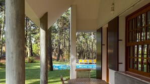 An outdoor patio with a wooden table and chairs, a concrete bench, and a view of a swimming pool and trees. - Garden, near the beach, car essential (Banzão)