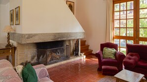 A living room with a fireplace, a red armchair, a wooden table, and a view of the outdoors through a large window. - Garden, near the beach, car essential (Banzão)