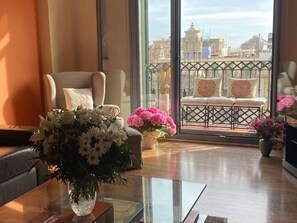 A room with a glass table, a chair, a sofa with cushions, and a balcony with a view of buildings.