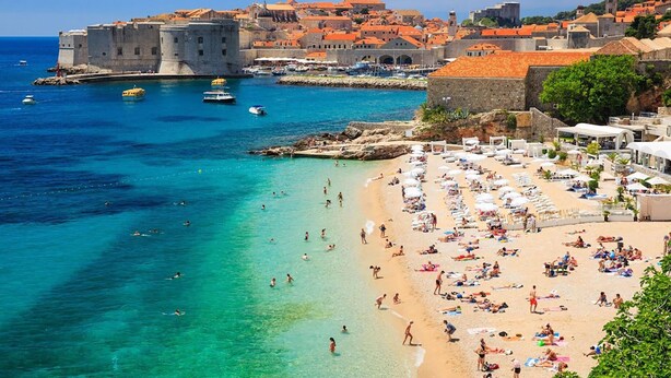 Un pueblo costero con una fortificación histórica, una playa de arena con sombrillas y aguas azul turquesa.