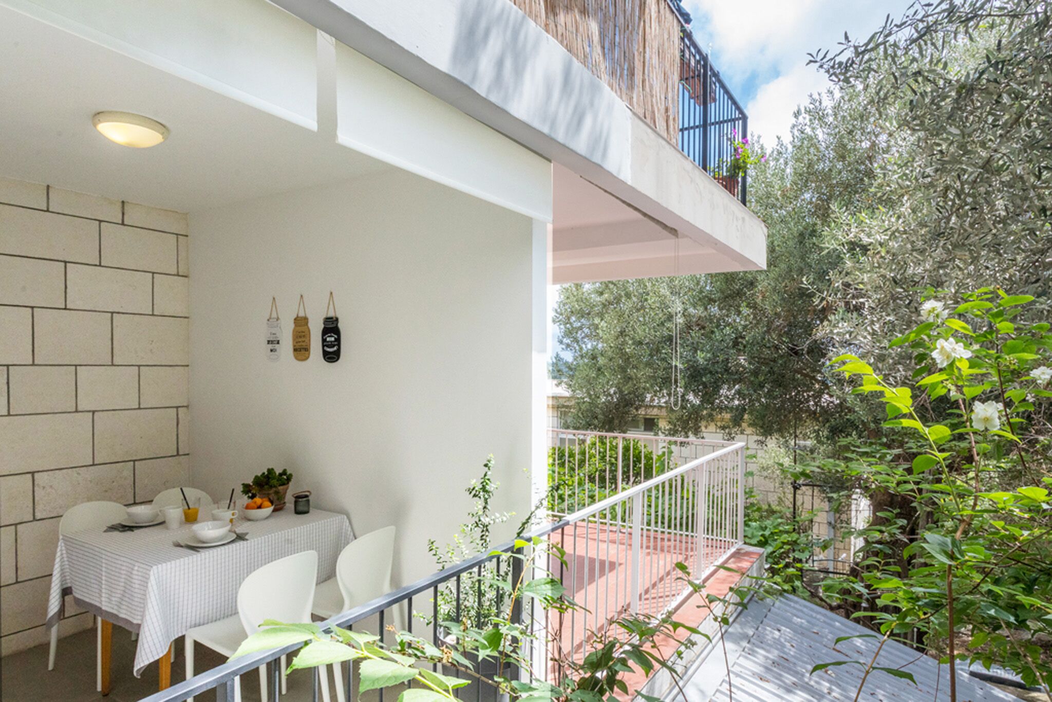 A dining area with a table set for four, a white wall, and a view of a garden with a metal railing.
