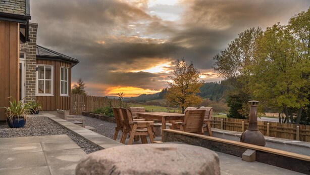 Une terrasse avec une table et des chaises en pierre, une clôture en bois et une vue sur un coucher de soleil se déclinant sur un paysage vallonné.