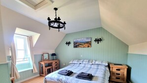 Une chambre à coucher avec un lit, des tables de chevet en bois, un bureau, un lustre, une fenêtre avec des rideaux et un tableau accroché au mur.