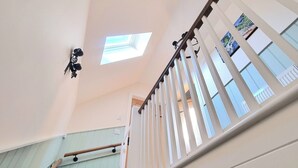 Un escalier avec une rampe en bois et des balustrades blanches, menant à un palier doté d’un puits de lumière et d’un luminaire fixé au mur.