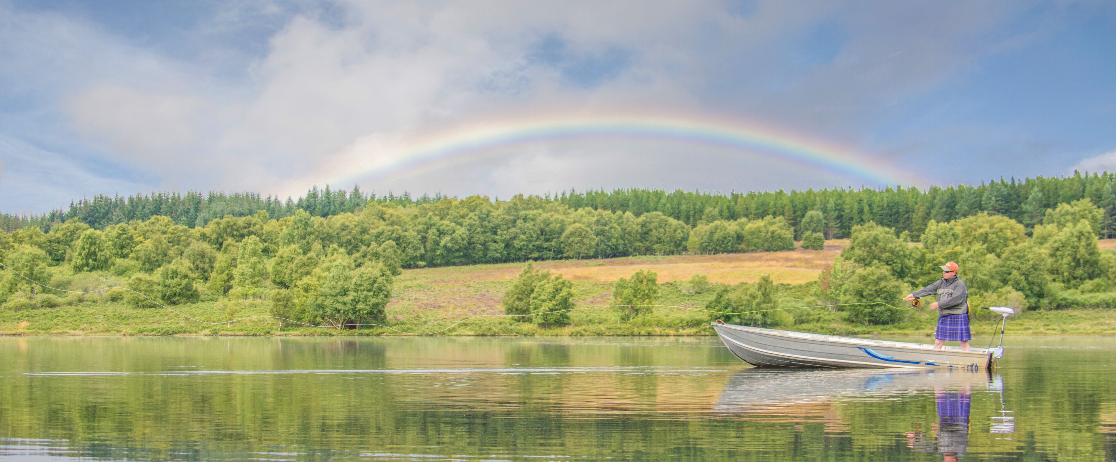A man fishing on a boat in a calm lake with a rainbow in the background.