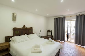 Quarto de hotel com uma cama grande, uma mesa de cabeceira de madeira, uma cadeira e uma varanda com cortinas.