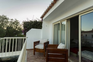 Uma varanda com duas cadeiras de madeira, uma parede branca e uma porta de vidro deslizante que dá para um espaço interior.