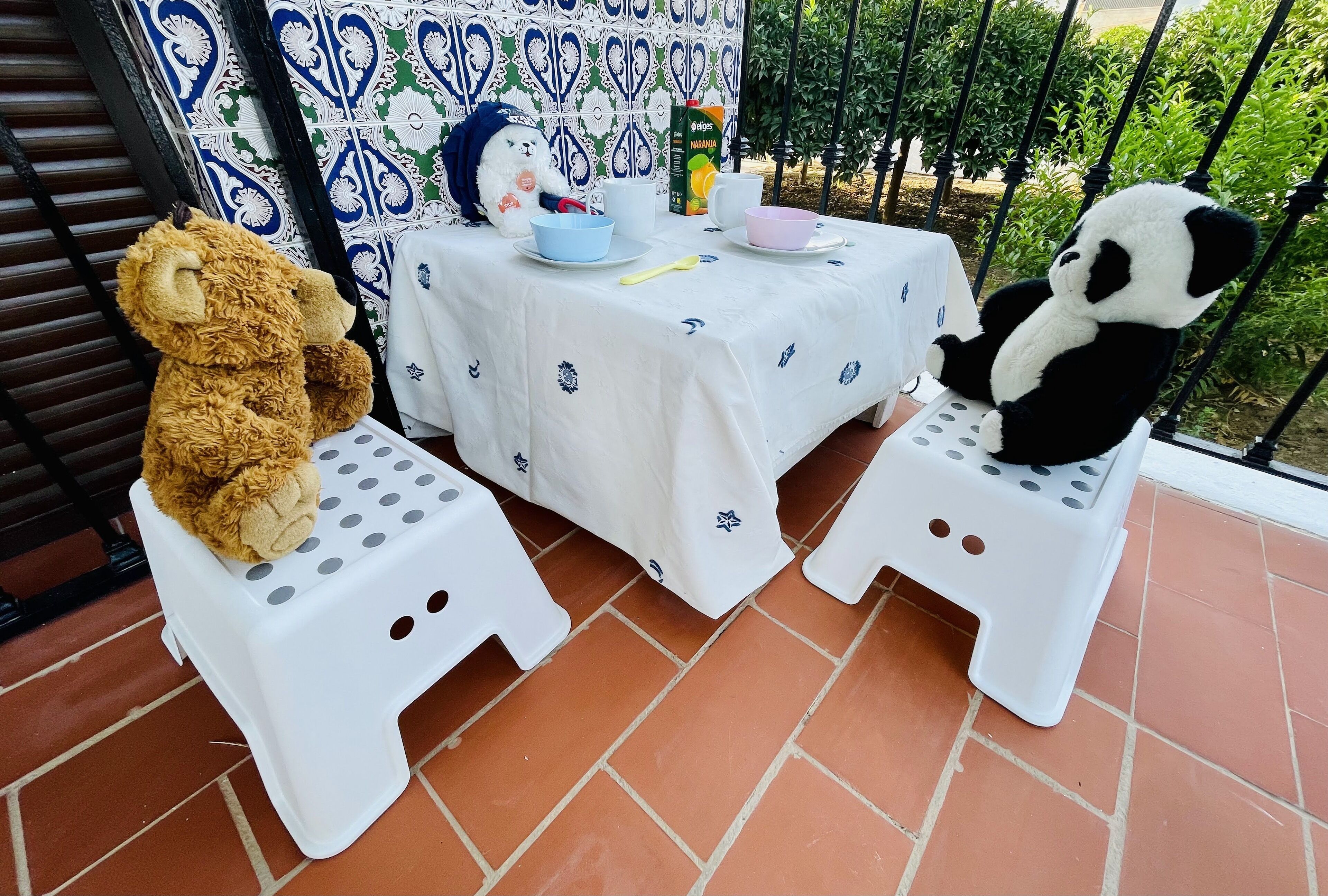 Two white plastic stools with a table between them, a teddy bear on the left stool, and a panda bear on the right.