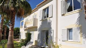 Ein zweistöckiges Gebäude mit weißen Wänden und Fensterläden, einem Balkon und einem kleinen Garten mit einer Palme und Liegestühlen.