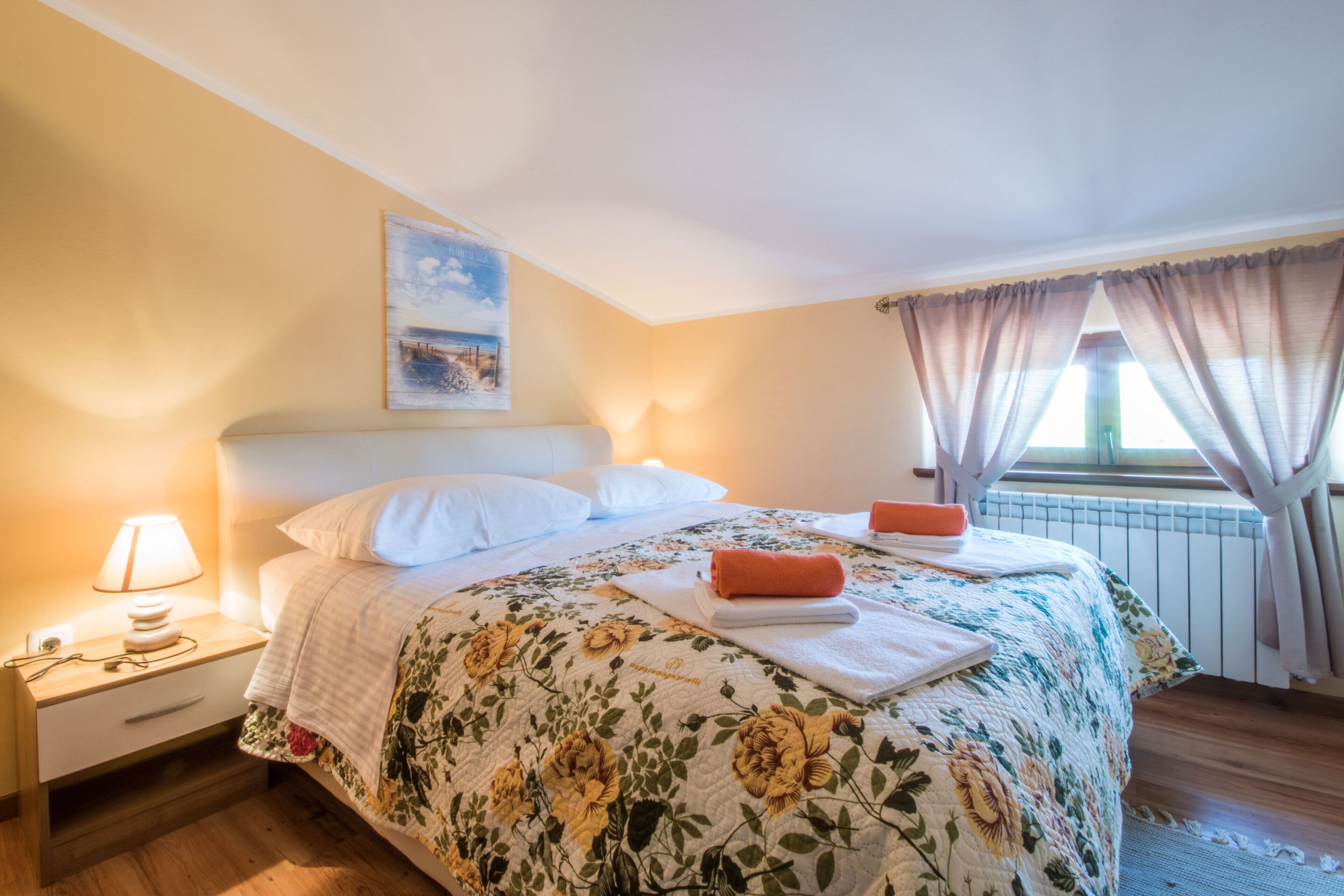 A neatly made bed with a floral bedspread, a nightstand with a lamp, and a window with curtains.