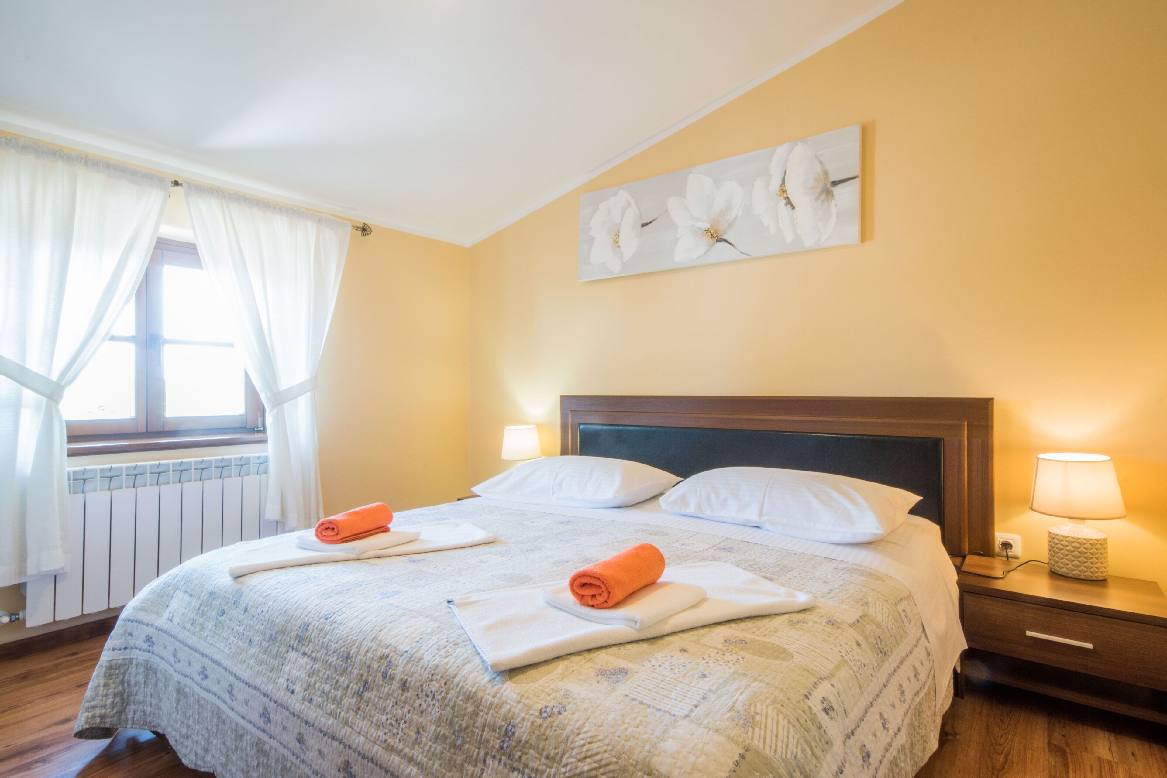 A neatly made bed with white linens and two rolled towels, a bedside table with a lamp, a window with white curtains, and a wall-mounted artwork of white flowers.