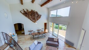 A spacious living area with a high ceiling, a dining set, and a large window providing a view of the outdoors. - Large Villa with heated pool and stunning views set among olive trees (Callas)