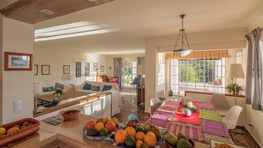 Ampla sala de estar com área de refeições, bancada na cozinha com frutas e nozes, e vista para o verde lá fora.