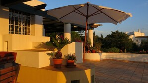 Uma área de pátio externa com mesa, cadeiras, plantas em vasos e um guarda-sol grande.