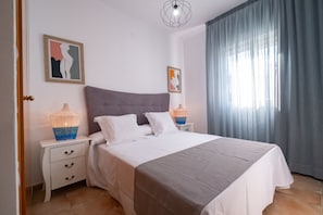 Ein ordentlich bezogenes Bett mit grauem Kopfteil, weißer Bettwäsche und zwei Nachttischlampen mit Flechtenschirmen.