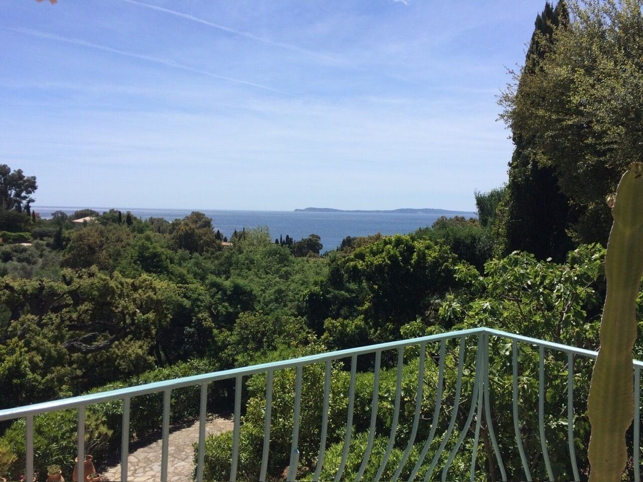 A balcony with a metal railing overlooking a lush garden and a distant view of the sea.