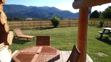 Une terrasse en bois avec une table et deux chaises, donnant sur une cour clôturée et offrant une vue sur les montagnes en arrière-plan.
