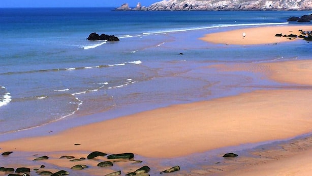 A beach with clear blue water, a sandy shore, and rocky outcrops in the distance.