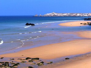 A beach with clear blue water, a sandy shore, and rocky outcrops in the distance.
