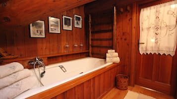 A wooden cabin bathroom with a bathtub, framed pictures on the wall, and a wooden door with lace curtains.