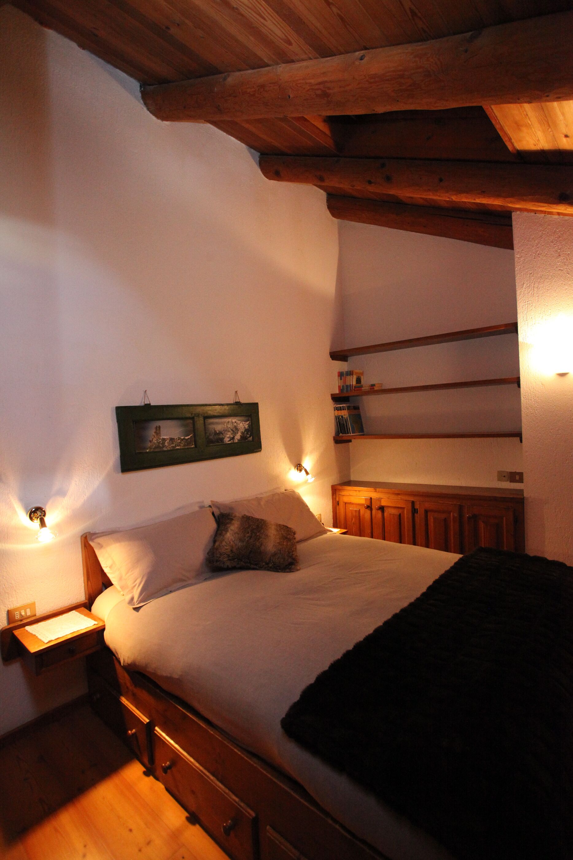 A bedroom with a wooden bed, a nightstand, a shelf with books, and a wall-mounted picture frame.