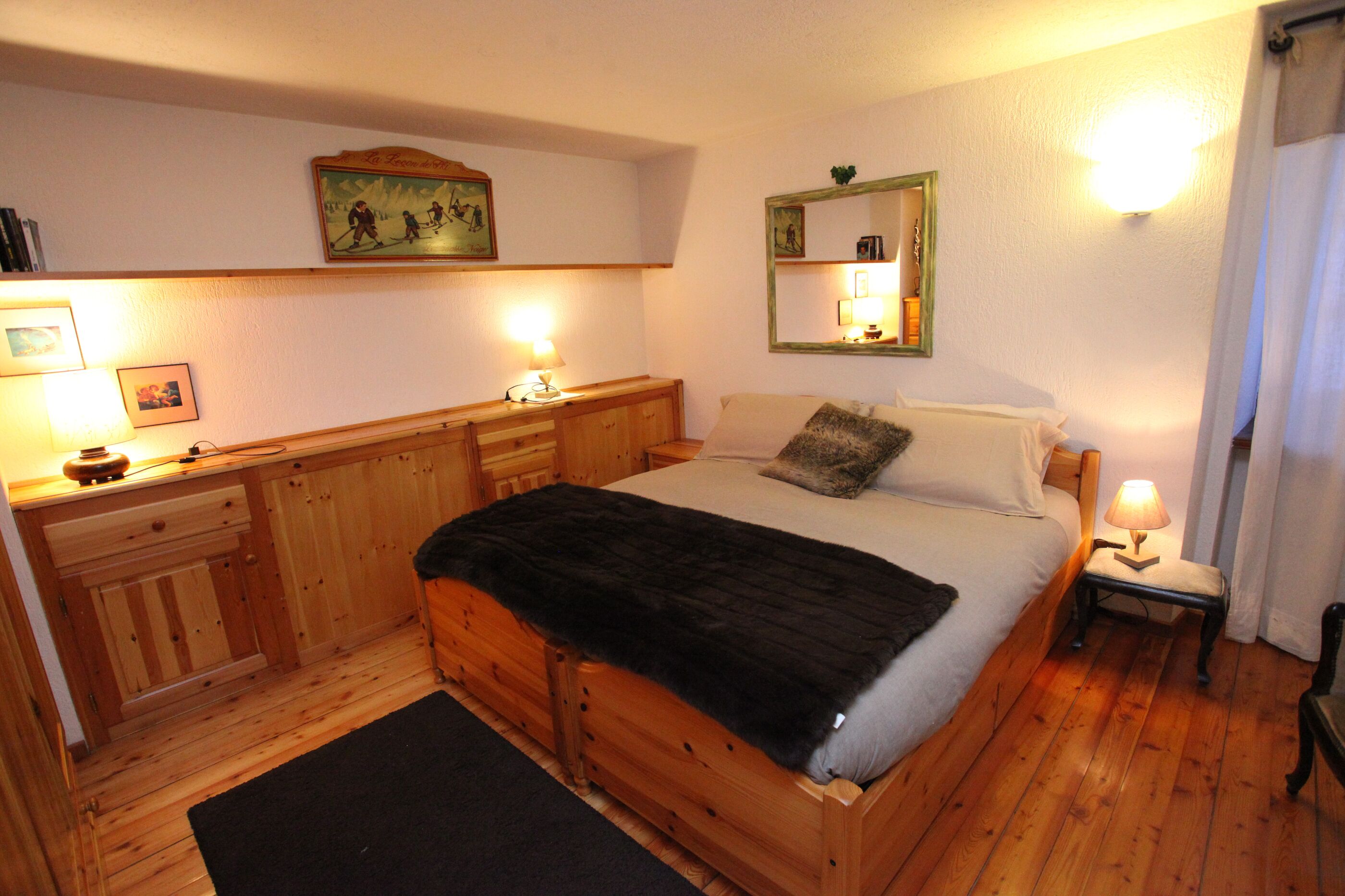 Een houten bed met een zwart deken, een nachtkastje met een lamp en een spiegel aan de muur.