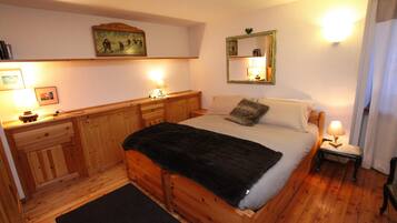 Een houten bed met een zwart deken, een nachtkastje met een lamp en een spiegel aan de muur.