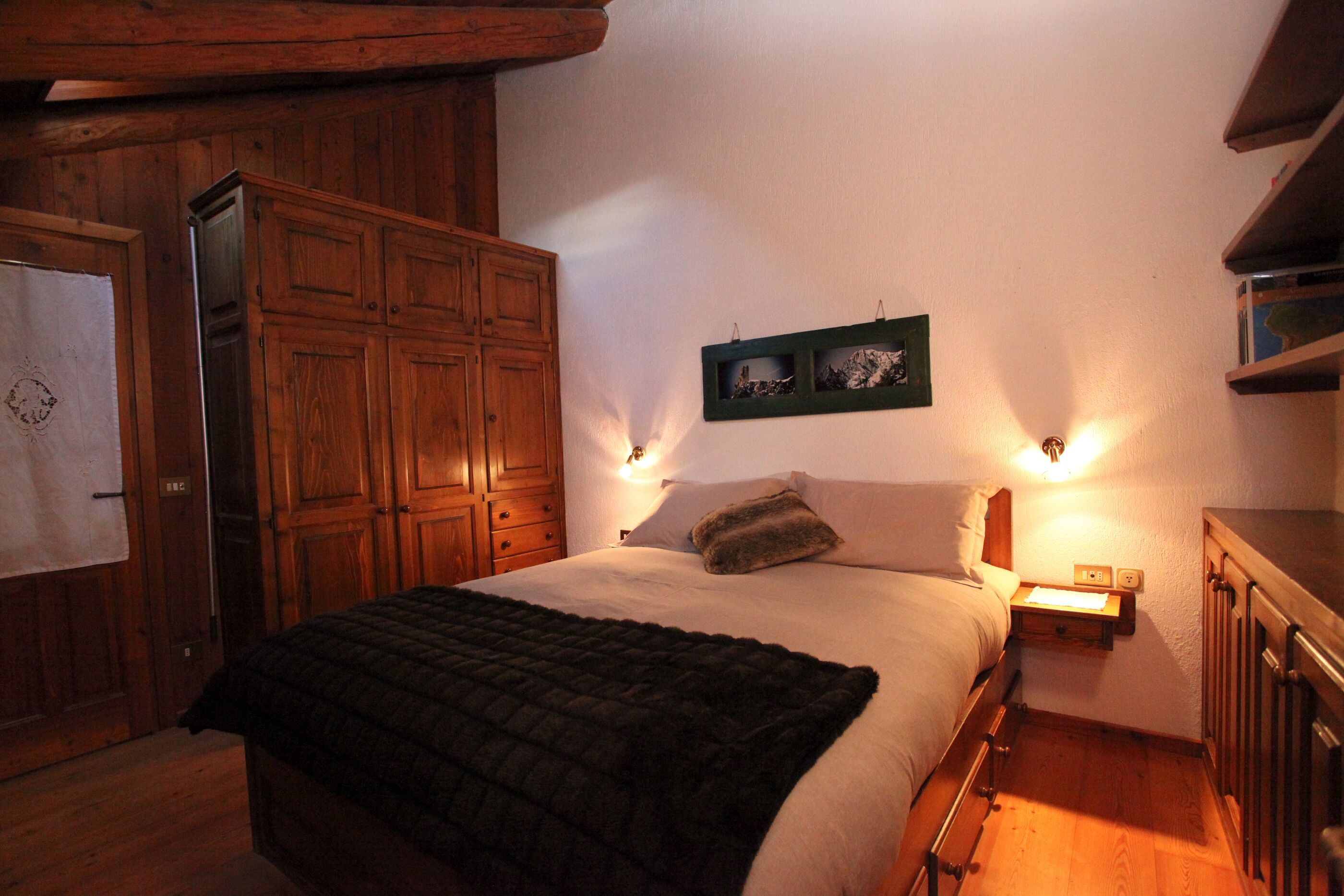 A bedroom with a wooden wardrobe, a bed with a dark blanket, two wall-mounted reading lamps, and a small shelf with books.