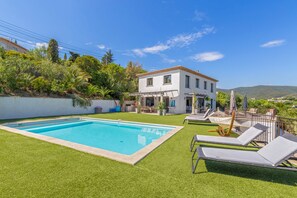 A pool with a white house in the background, surrounded by greenery and a clear sky.