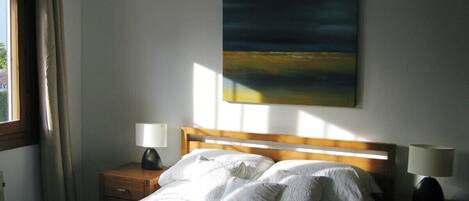 Un dormitorio con cama, dos mesitas de noche con lámparas, una ventana con cortinas y un cuadro en la pared.