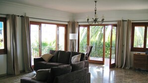 Ein geräumiges Wohnzimmer mit einem dunkelbraunen Ecksofa, einem kleinen Hocker und einer Stehlampe. Große Schiebegläser führen zu einem Balkon mit Blick auf Palmen.