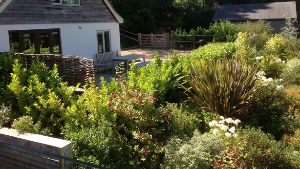 Ein Haus mit schrägem Dach, einer Holzdiele und einem Garten mit verschiedenen Pflanzen und Blumen.