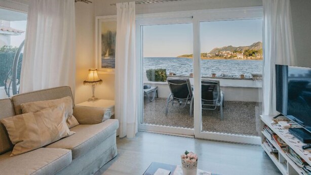 A living room with a sofa, a TV, and a table with chairs. There is a large window with a view of the sea and buildings. - TRANQUILITY, REST AND RELAXATION BY THE SEA.  IN WINTER CENTRAL HEATING  (Alcudia)