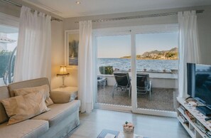 Een woonkamer met een bank, een televisie en een tafel met stoelen. Er is een groot raam met uitzicht op zee en gebouwen.