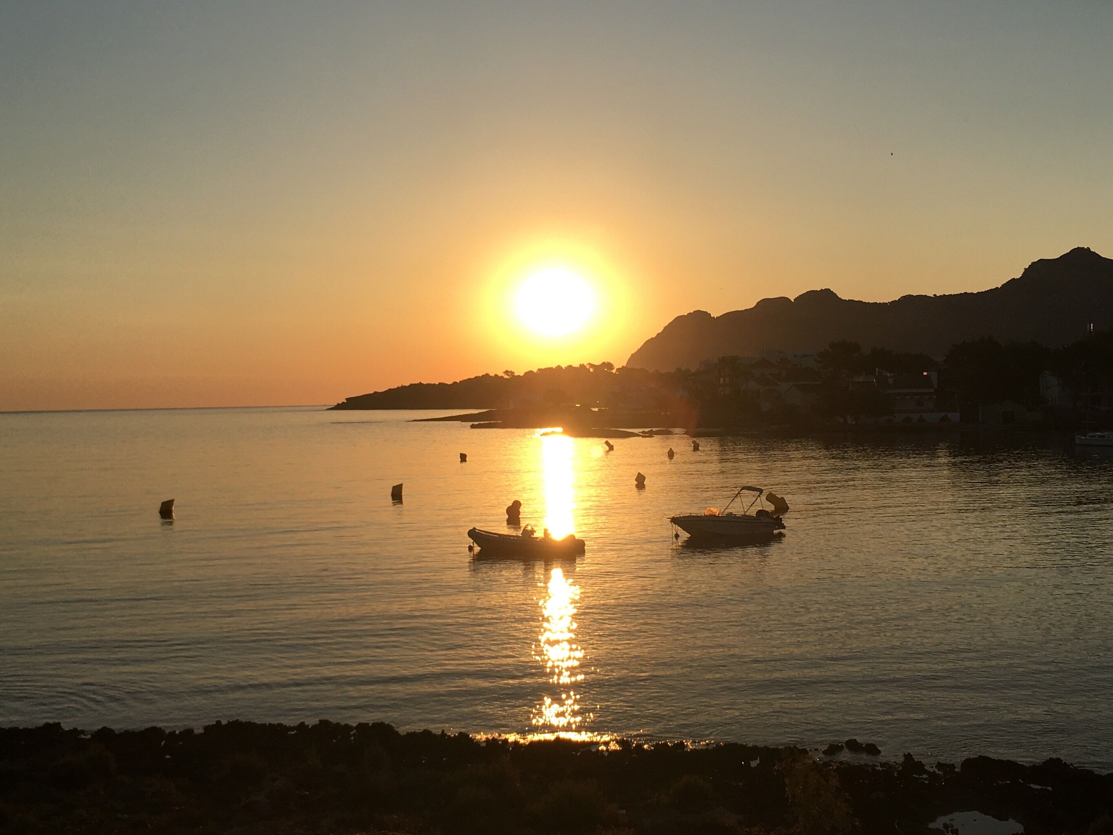 Un coucher de soleil sur une mer calme, avec des bateaux et une côte lointaine.