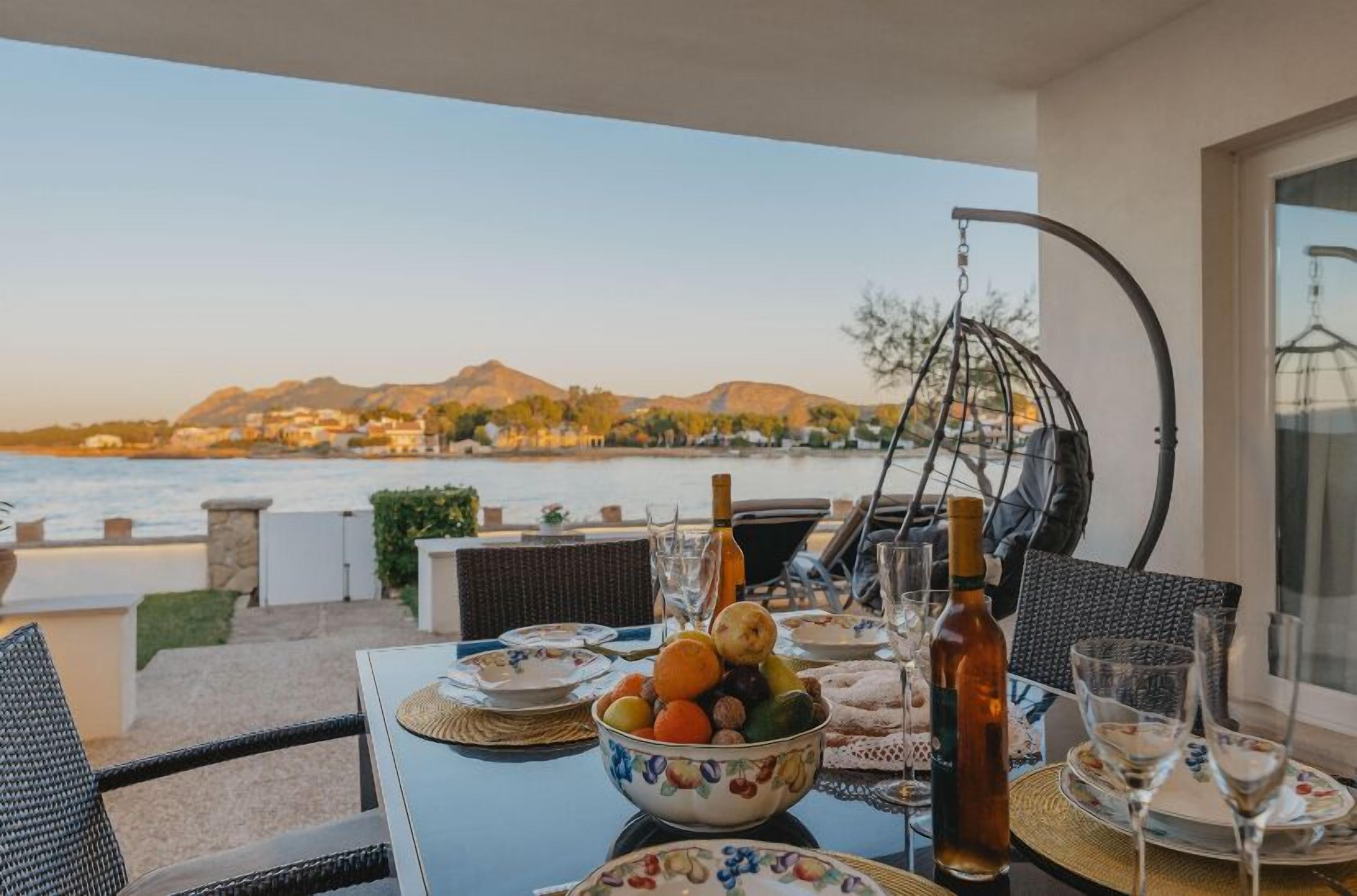 A dining table set with a fruit bowl, wine glasses, and bottles, overlooking a coastal view with mountains in the background.